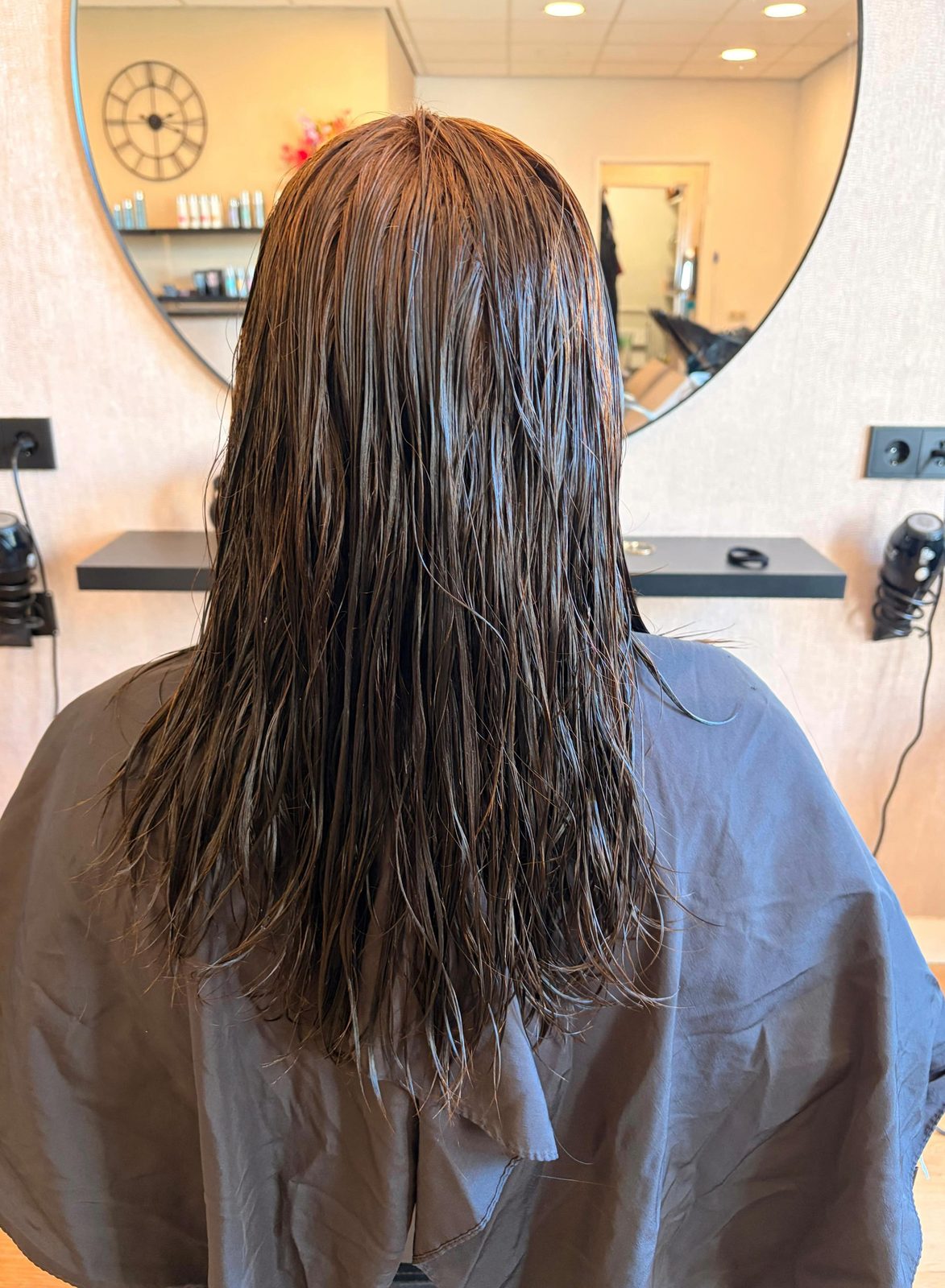 De salon in de spiegel: wandklok, werkplekken en een klant met nat haar in de stoel